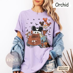 May include: A light purple t-shirt featuring a cartoon image of Mater from the movie Cars, decorated for Halloween. The design includes Mickey Mouse-shaped balloons, bats, ghosts, and a jack-o'-lantern. The word "Orchid" is printed on the top right.