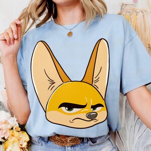May include: Light blue t-shirt featuring a cartoon illustration of a fennec fox's face. The fox has large ears, green eyes, and a slightly grumpy expression. The shirt is a solid color and appears to be made of a soft material.