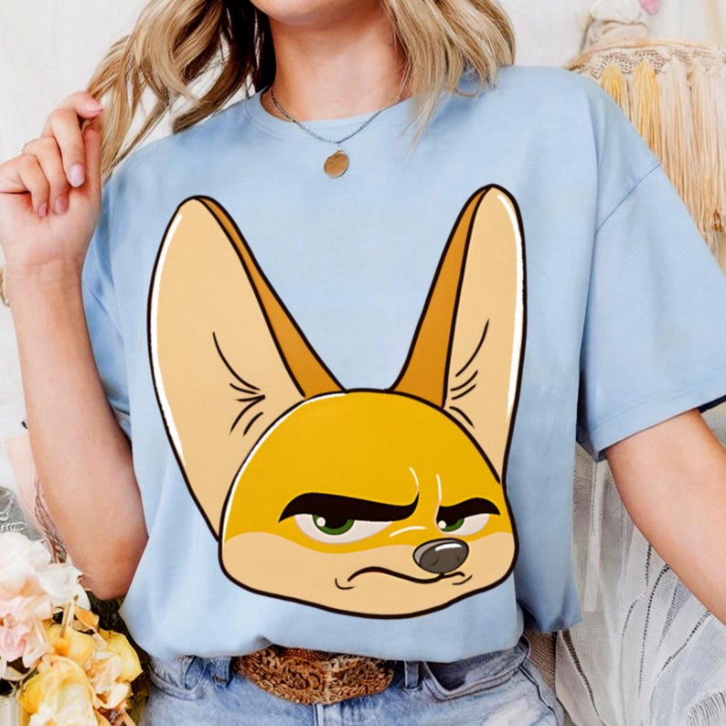 May include: Light blue t-shirt featuring a cartoon illustration of a fennec fox's face. The fox has large ears, green eyes, and a slightly grumpy expression. The shirt is a solid color and appears to be made of a soft material.