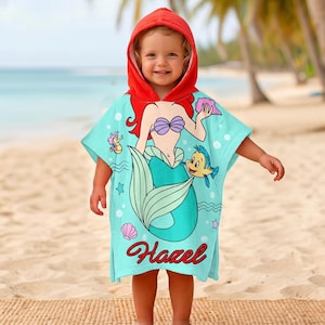 May include: A hooded turquoise beach towel featuring a mermaid design with red hair, a purple seashell bra, and a teal tail. The towel has a red hood and the name "Hazel" in cursive. The design includes a seahorse and a yellow fish.
