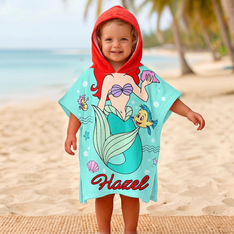 May include: A hooded turquoise beach towel featuring a mermaid design with red hair, a purple seashell bra, and a teal tail. The towel has a red hood and the name "Hazel" in cursive. The design includes a seahorse and a yellow fish.