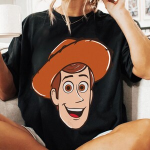 May include: Black t-shirt featuring a cartoon illustration of a cowboy character with a brown hat, brown hair, and a smiling face. The character's face is outlined in white. The shirt is made of a soft material.