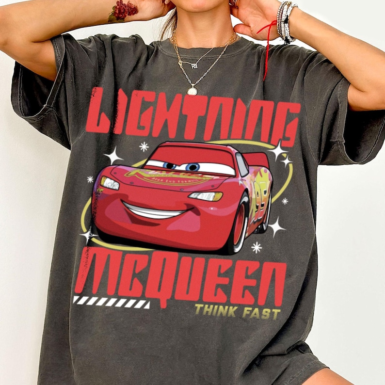 May include: A charcoal gray short-sleeve t-shirt featuring a graphic of a red race car. The text "LIGHTNING MCQUEEN" appears in large red letters above and below the car, with the words "THINK FAST" printed in yellow at the bottom.