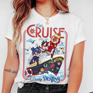 May include: White t-shirt featuring a colorful graphic of a cruise ship with cartoon characters. The design includes the text "Let's Cruise" and "Disney Destiny". The ship has a red and blue border with a nautical theme.