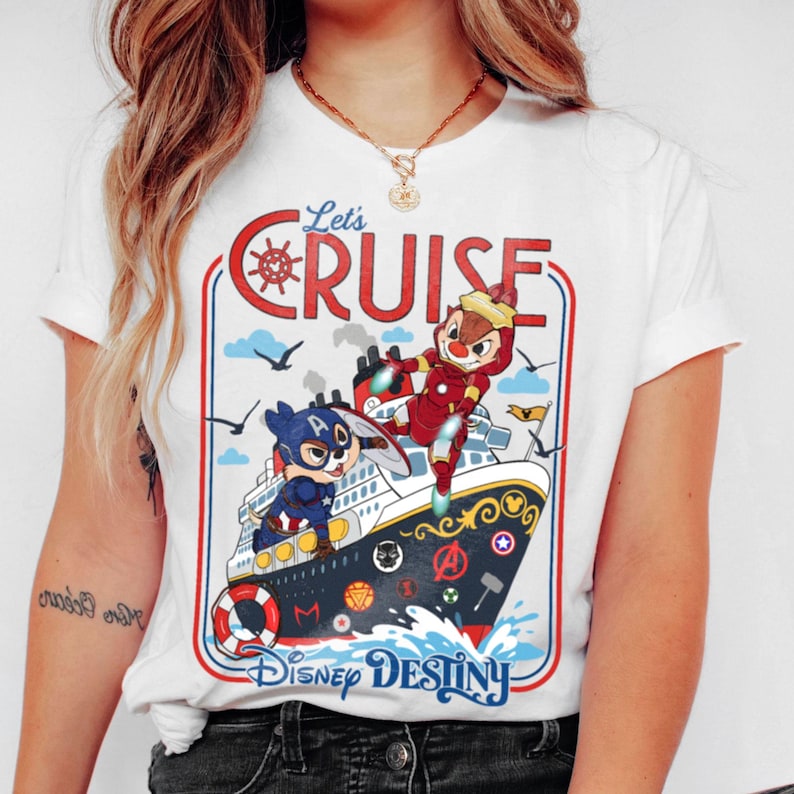 May include: White t-shirt featuring a colorful graphic of a cruise ship with cartoon characters. The design includes the text "Let's Cruise" and "Disney Destiny". The ship has a red and blue border with a nautical theme.