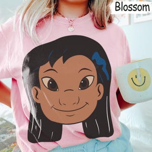 Puede incluir: Camiseta rosa claro con un dibujo animado de una ni&ntilde;a sonriente con cabello oscuro y piel morena. La palabra "Blossom" est&aacute; impresa en blanco. Una taza azul claro y blanca con una carita sonriente amarilla se sostiene en la mano derecha.