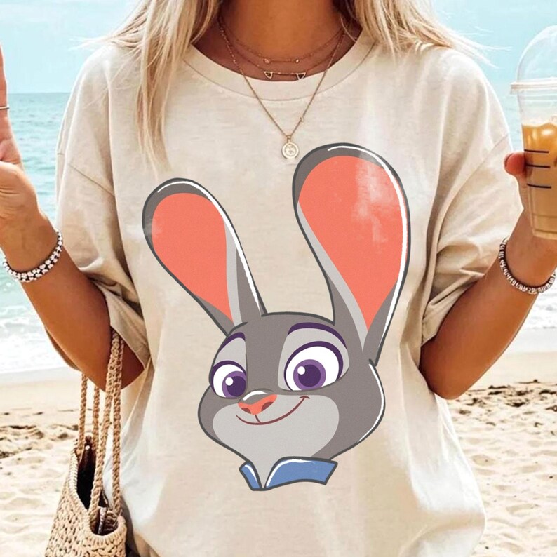 Puede incluir: Camiseta beige con una imagen de dibujos animados de un conejo gris con orejas rosas, ojos morados y un cuello azul. El conejo est&aacute; sonriendo. La camiseta la lleva una persona en la playa.