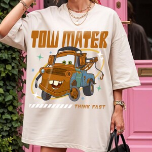 May include: A cream-colored short-sleeve t-shirt featuring a graphic of the character Tow Mater from the movie Cars. The design includes the text TOW MATER in large brown letters above the truck and the text THINK FAST in smaller brown letters.