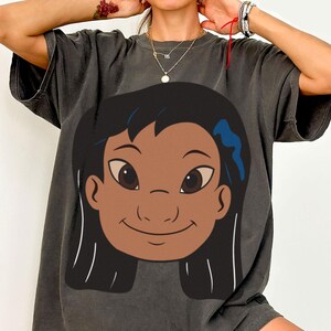 Puede incluir: Camiseta gris oscuro con una ilustraci&oacute;n de dibujos animados de una ni&ntilde;a con cabello negro, ojos marrones y un accesorio azul para el cabello. El gr&aacute;fico est&aacute; centrado en la parte delantera de la camiseta.