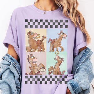 May include: A light purple t-shirt featuring a cartoon horse design in four panels. The horse is brown with a saddle and a smiling expression. The design is framed by a black and white checkered pattern at the top and bottom.