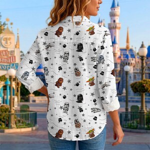 May include: White button-down shirt with a Star Wars character pattern. The shirt has long sleeves and a classic collar. The pattern includes characters like Darth Vader, Baby Yoda, and Stormtroopers, along with stars and spaceships.