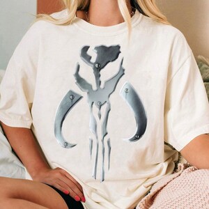 May include: Off-white t-shirt with a metallic silver emblem. The emblem is a symmetrical design with curved and angular elements, resembling a helmet or mask. The shirt is made of a soft material.