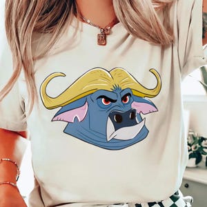 May include: A white t-shirt featuring a cartoon buffalo head. The buffalo has a blue face, yellow horns, and red eyes. The design is centered on the shirt, creating a bold graphic.