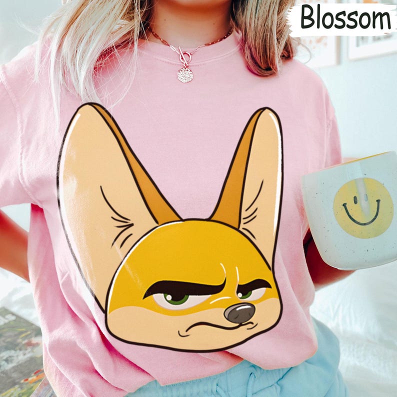 May include: Pink sweatshirt featuring a large, cartoonish illustration of a fennec fox's face. The fox has large ears, a yellow and tan color scheme, and a grumpy expression. The word "Blossom" is printed in white in the upper right corner.