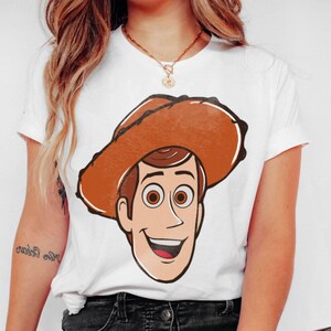 May include: White t-shirt featuring a cartoon illustration of a smiling cowboy character with a brown hat. The character has brown hair and eyes, and a tan face. The shirt is a classic crew neck style.