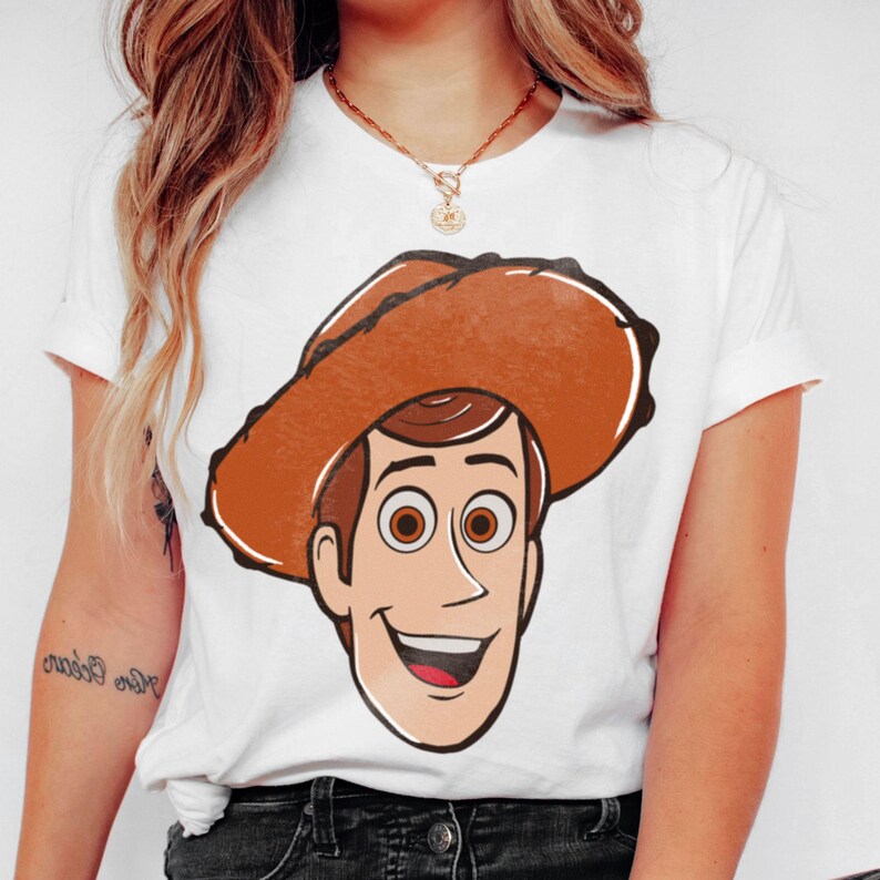 May include: White t-shirt featuring a cartoon illustration of a smiling cowboy character with a brown hat. The character has brown hair and eyes, and a tan face. The shirt is a classic crew neck style.
