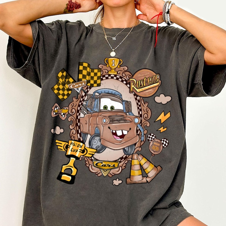 May include: Dark gray t-shirt featuring a cartoon image of Mater from the movie Cars. The design includes a checkered flag, trophy, and the text "Rusteze" and "Cars". The shirt has a vintage, distressed look.