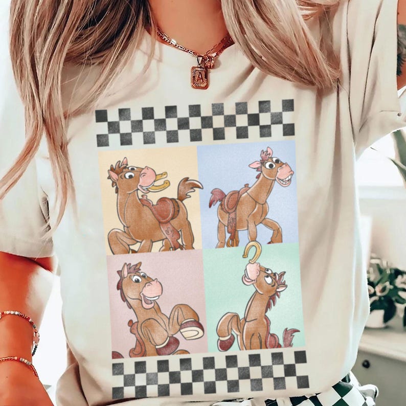 May include: A cream-colored t-shirt featuring a cartoon graphic of a brown horse in four different poses. The graphic is set against a backdrop of pastel colors and a black and white checkered pattern.