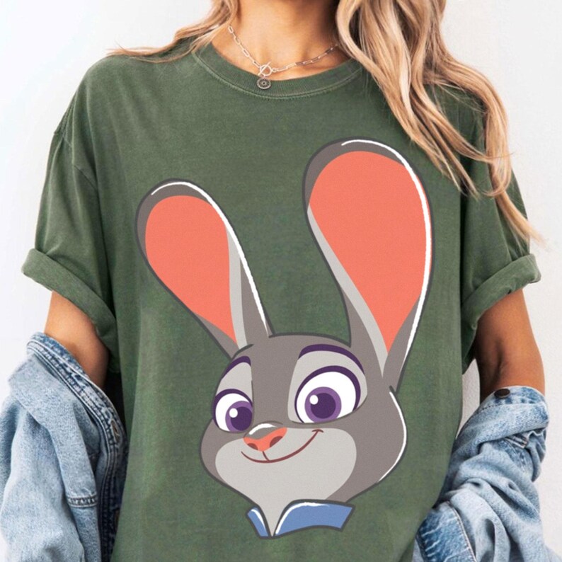 Puede incluir: Camiseta verde oliva con un dise&ntilde;o de conejo de dibujos animados. El conejo es gris con orejas naranjas, ojos morados y un cuello azul. La camiseta tiene las mangas enrolladas y se lleva con una chaqueta vaquera.