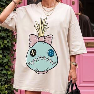 May include: Oversized beige t-shirt featuring a cartoon voodoo doll design. The doll has a light blue face with a pink bow, yellow hair, and stitched details. The shirt has short sleeves and a relaxed fit.