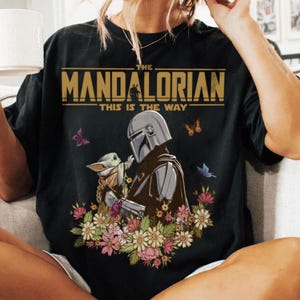 May include: Black t-shirt featuring a graphic of The Mandalorian and Grogu surrounded by flowers and butterflies. The text "THE MANDALORIAN" and "THIS IS THE WAY" are printed in gold.