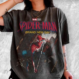 May include: Dark gray t-shirt featuring the Marvel Studios Spider-Man logo in red and blue, with the words "BRAND NEW DAY." The graphic shows Spider-Man in action against a cityscape backdrop. The shirt has a vintage, worn look.