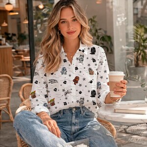 May include: White button-down shirt with a Star Wars design. The shirt is decorated with various characters, starships, and star graphics. The shirt is worn with blue jeans.