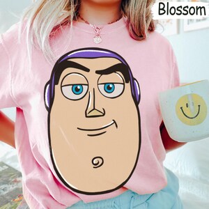 May include: Pink sweatshirt featuring a large cartoon face with blue eyes, purple accents, and a smiling expression. The sweatshirt is a light pink color. A white mug with a yellow smiley face is held in the image.
