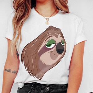 May include: White t-shirt featuring a cartoon sloth face with brown fur, green eyes, and a black nose. The sloth is smiling. The shirt has short sleeves and a crew neck.