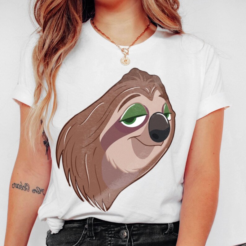 May include: White t-shirt featuring a cartoon sloth face with brown fur, green eyes, and a black nose. The sloth is smiling. The shirt has short sleeves and a crew neck.