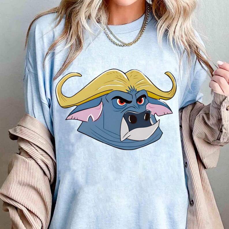 May include: Light blue t-shirt featuring a cartoon buffalo head. The buffalo has a blue face, yellow horns, red eyes, and a white tooth. The shirt is a solid light blue color.