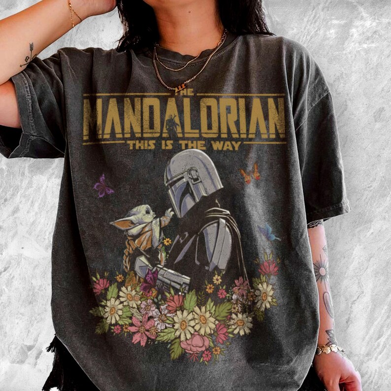 May include: Dark gray t-shirt featuring a graphic of The Mandalorian and Grogu surrounded by flowers and butterflies. The text "THE MANDALORIAN THIS IS THE WAY" is printed across the top in gold.