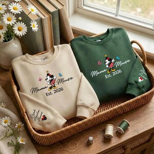 K&ouml;nnte beinhalten: Zwei Sweatshirts, eines beige und eines gr&uuml;n, werden in einem Weidenkorb pr&auml;sentiert. Beide zeigen ein Minnie Mouse-Design mit dem Text "Mama Mouse Est. 2026". Das beige Sweatshirt hat die Namen "Elijah Amelia James" auf dem &Auml;rmel.