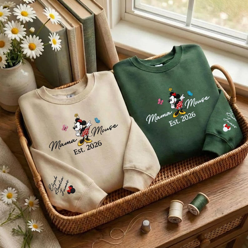 K&ouml;nnte beinhalten: Zwei Sweatshirts, eines beige und eines gr&uuml;n, werden in einem Weidenkorb pr&auml;sentiert. Beide zeigen ein Minnie Mouse-Design mit dem Text "Mama Mouse Est. 2026". Das beige Sweatshirt hat die Namen "Elijah Amelia James" auf dem &Auml;rmel.