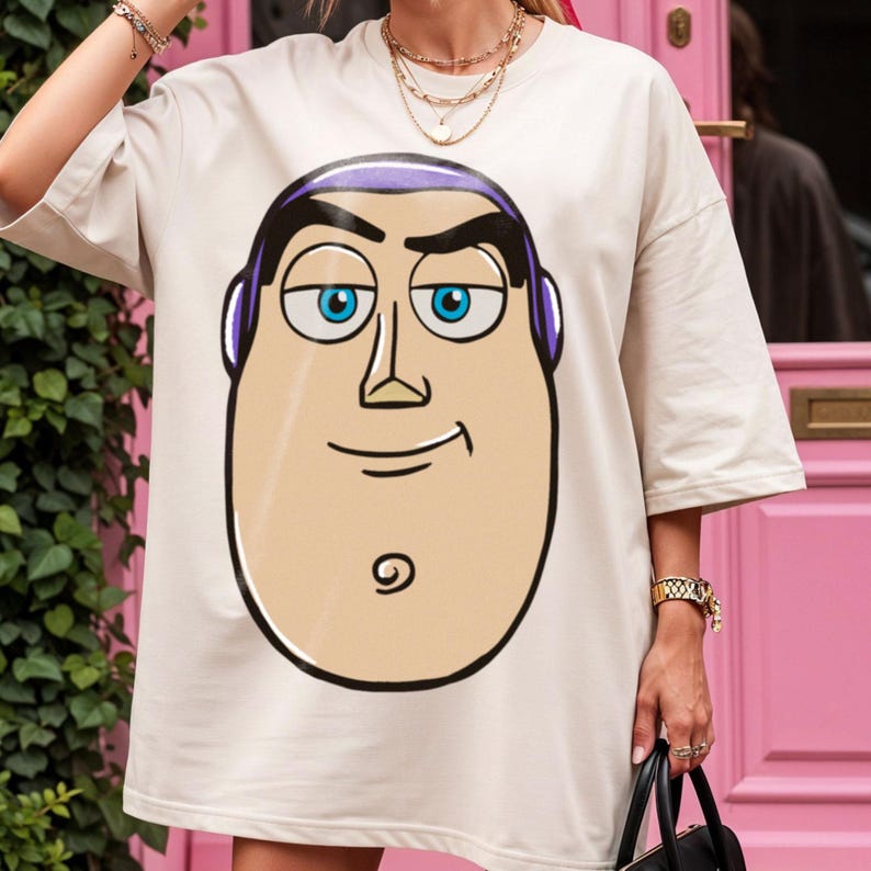 May include: Oversized, cream-colored t-shirt featuring a large cartoon face with blue eyes, purple hair, and a black outline. The design is centered on the front of the shirt. The shirt has short sleeves and a relaxed fit.