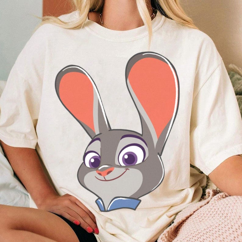 Puede incluir: Camiseta color crema con una ilustraci&oacute;n de dibujos animados de Judy Hopps de Zootopia. El gr&aacute;fico muestra la cara de Judy con pelaje gris, orejas grandes con forro rosa y ojos morados. La camiseta tiene un corte holgado.