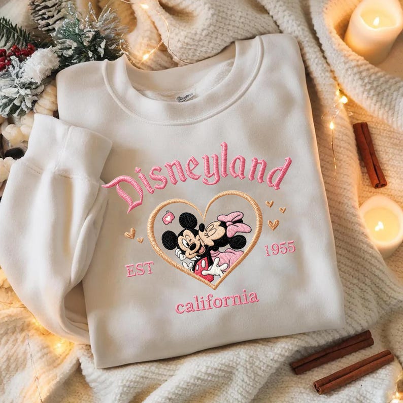 Peut inclure: Un sweat-shirt blanc avec une broderie rose "Disneyland". Le motif comprend Mickey et Minnie Mouse dans un c&oelig;ur, avec "EST 1955" et "california" en dessous. Le sweat-shirt a un col rond et des manches longues.