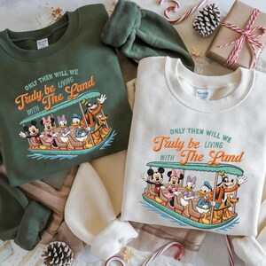 May include: Two sweatshirts, one green and one cream, featuring a colorful embroidered design of Disney characters on a boat. The text "ONLY THEN WILL WE Truly be LIVING WITH The Land" is above the image. Festive holiday decor surrounds the sweatshirts.