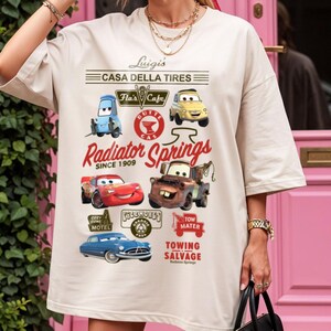Pu&ograve; includere: Una t-shirt beige con una stampa grafica di personaggi e loghi del film Cars. Il design include testi come Luigi's Casa Della Tires, Radiator Springs Since 1909 e Tow Mater Towing Salvage, insieme a illustrazioni di vari veicoli.