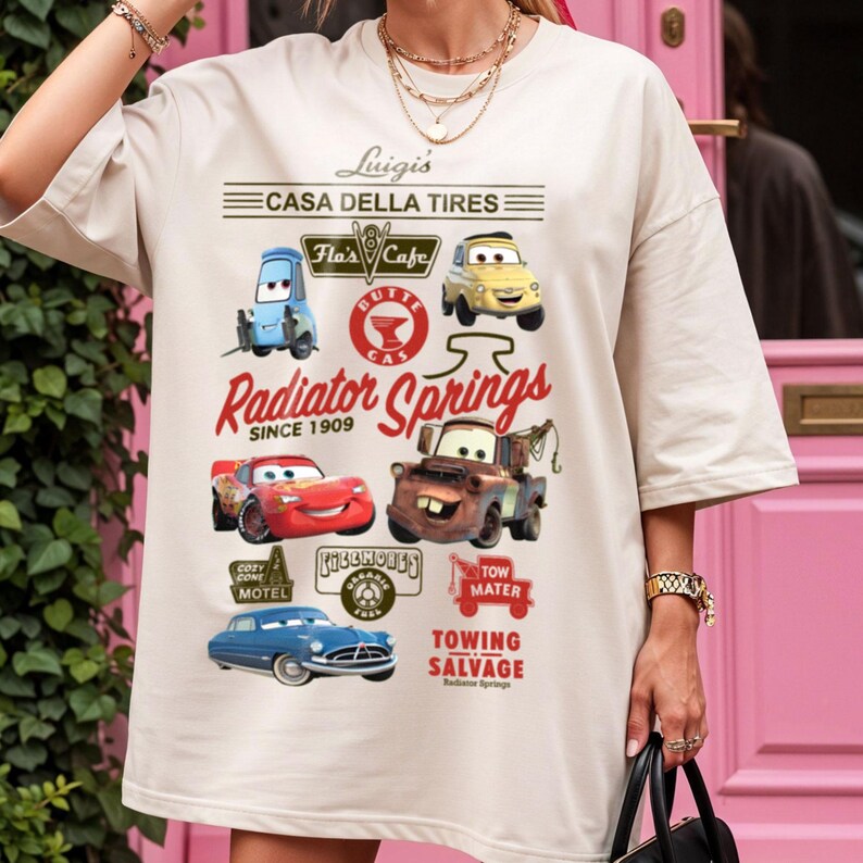 Pu&ograve; includere: Una t-shirt beige con una stampa grafica di personaggi e loghi del film Cars. Il design include testi come Luigi's Casa Della Tires, Radiator Springs Since 1909 e Tow Mater Towing Salvage, insieme a illustrazioni di vari veicoli.