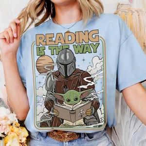 Puede incluir: Camiseta azul claro con un dise&ntilde;o de The Mandalorian y Grogu leyendo un libro, con el texto "READING IS THE WAY" en estilo retro. El dise&ntilde;o incluye un planeta y nubes, creando una escena del universo Star Wars.