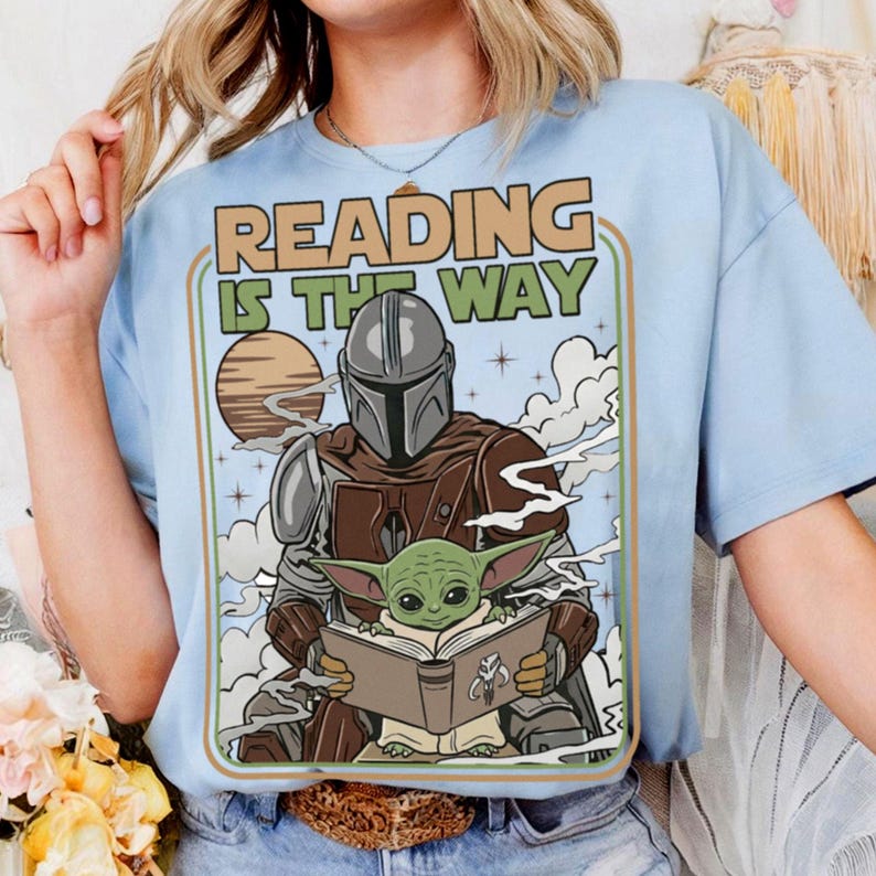Puede incluir: Camiseta azul claro con un dise&ntilde;o de The Mandalorian y Grogu leyendo un libro, con el texto "READING IS THE WAY" en estilo retro. El dise&ntilde;o incluye un planeta y nubes, creando una escena del universo Star Wars.