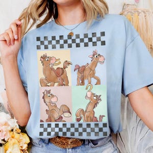 May include: Light blue t-shirt featuring a cartoon horse design in four panels. The horse is brown with a pink mane and a saddle. The shirt has a checkered pattern at the top and bottom. The shirt is made of a soft material.