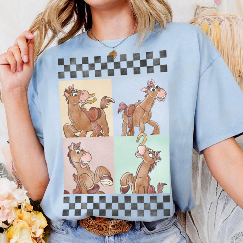 May include: Light blue t-shirt featuring a cartoon horse design in four panels. The horse is brown with a pink mane and a saddle. The shirt has a checkered pattern at the top and bottom. The shirt is made of a soft material.