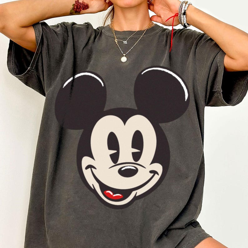 May include: Dark gray t-shirt featuring a large graphic of Mickey Mouse's face. The design includes black ears, a white face, and a smiling expression with a red mouth. The shirt has short sleeves and a relaxed fit.