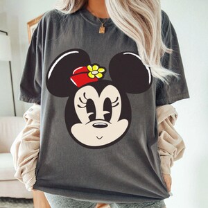 Peut inclure: T-shirt gris fonc&eacute; avec un grand dessin du visage de Minnie Mouse. Minnie Mouse est repr&eacute;sent&eacute;e avec des oreilles noires, un n&oelig;ud rouge avec une fleur jaune et une expression souriante. Le t-shirt est de style d&eacute;contract&eacute; et surdimensionn&eacute;.