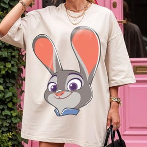 Puede incluir: Camiseta beige de gran tama&ntilde;o con una ilustraci&oacute;n de dibujos animados de Judy Hopps de Zootopia. El gr&aacute;fico muestra la cara de Judy con pelaje gris, orejas grandes con forro rosa y un cuello azul. La camiseta tiene mangas cortas.