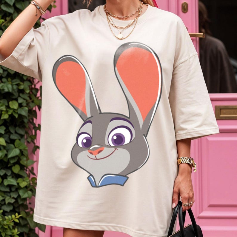 Puede incluir: Camiseta beige de gran tama&ntilde;o con una ilustraci&oacute;n de dibujos animados de Judy Hopps de Zootopia. El gr&aacute;fico muestra la cara de Judy con pelaje gris, orejas grandes con forro rosa y un cuello azul. La camiseta tiene mangas cortas.