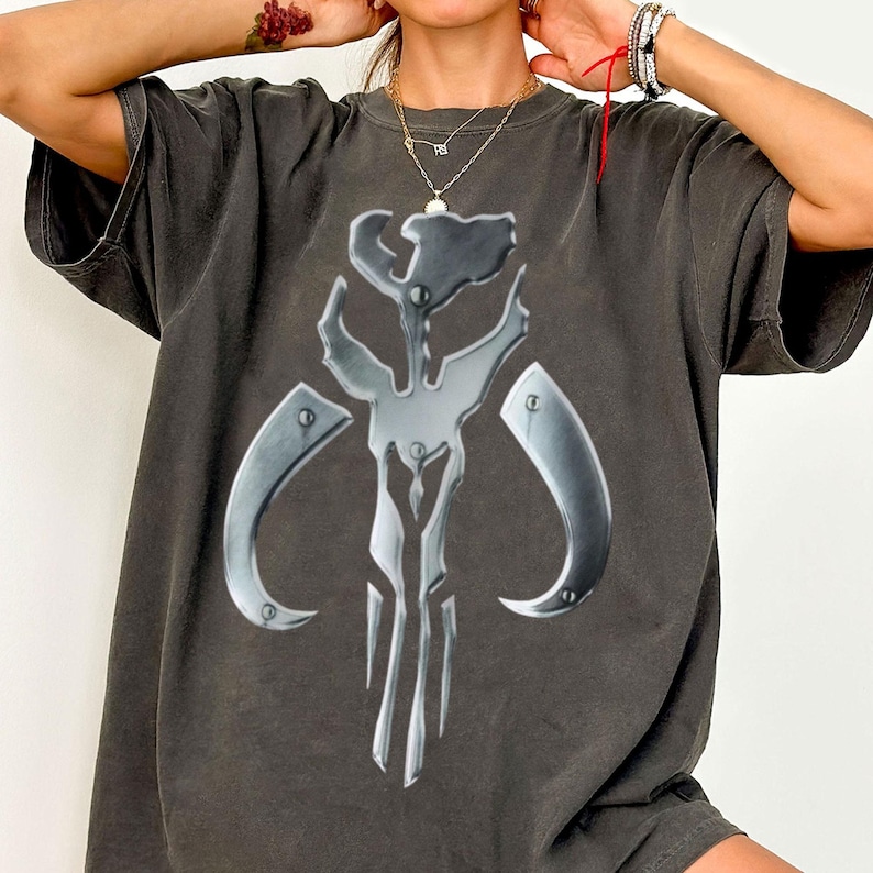 May include: Dark grey t-shirt with a metallic silver emblem. The emblem is a stylized skull with curved horns and a central vertical element. The shirt has a vintage, worn appearance.