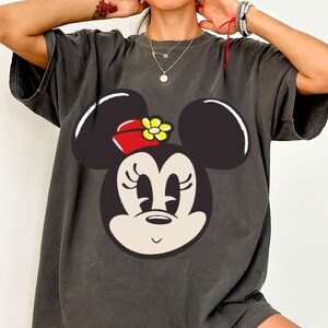 Peut inclure: T-shirt gris fonc&eacute; avec un grand motif de Minnie Mouse. Le dessin repr&eacute;sente le visage de Minnie avec des oreilles noires, un n&oelig;ud rouge et une fleur jaune. Le t-shirt a des manches courtes et une coupe d&eacute;contract&eacute;e, id&eacute;al pour un usage quotidien.